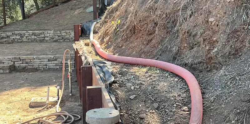 Red suction hose run down a hillside to an open septic riser beside a retaining wall