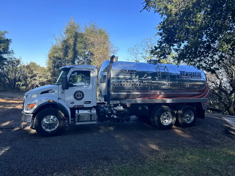 Ryan's polished stainless vacuum truck parked on a gravel drive under oak trees